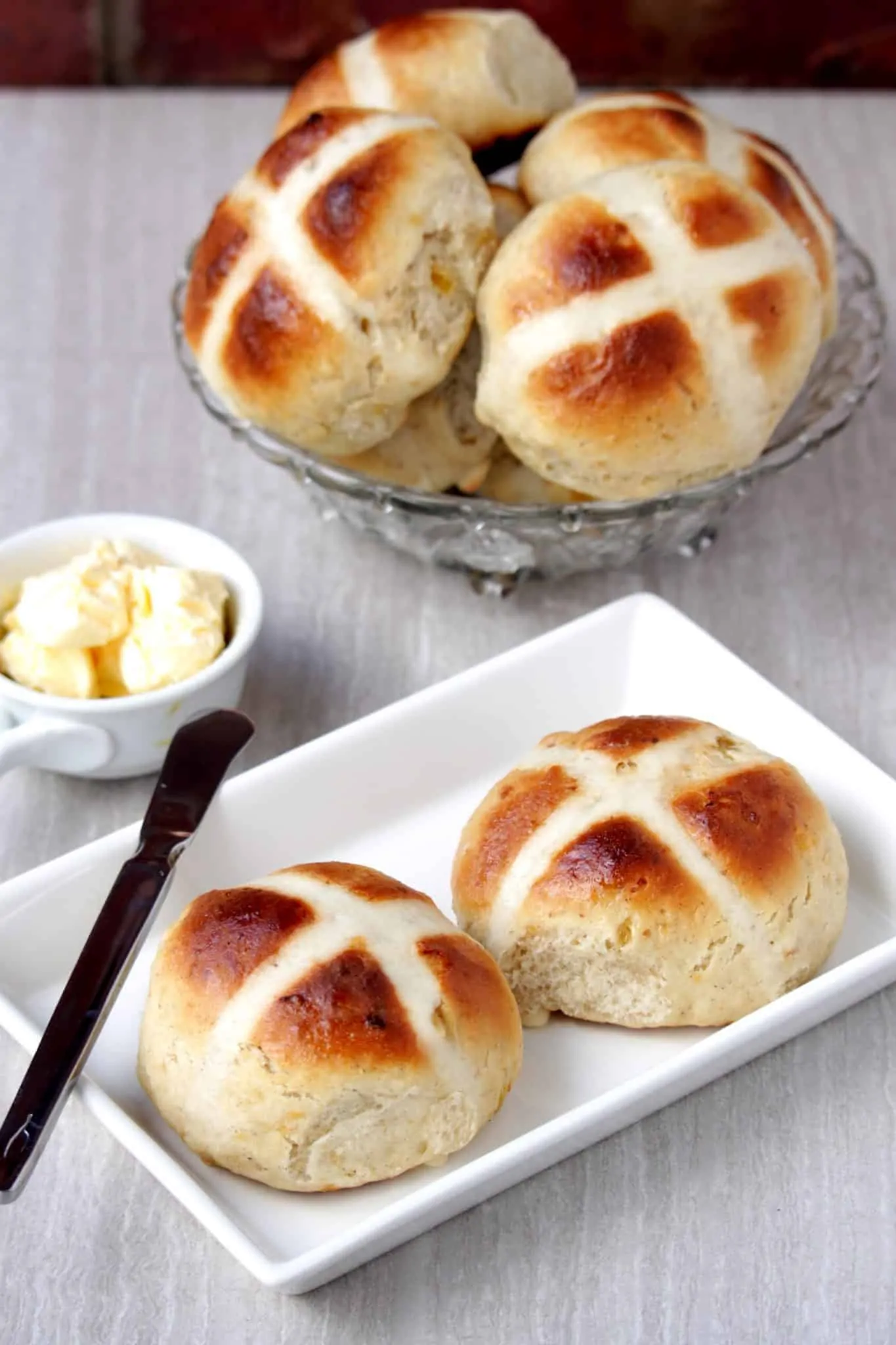 Hot Cross Buns in a white plate and stacked in a glass bowl with butter on the side.