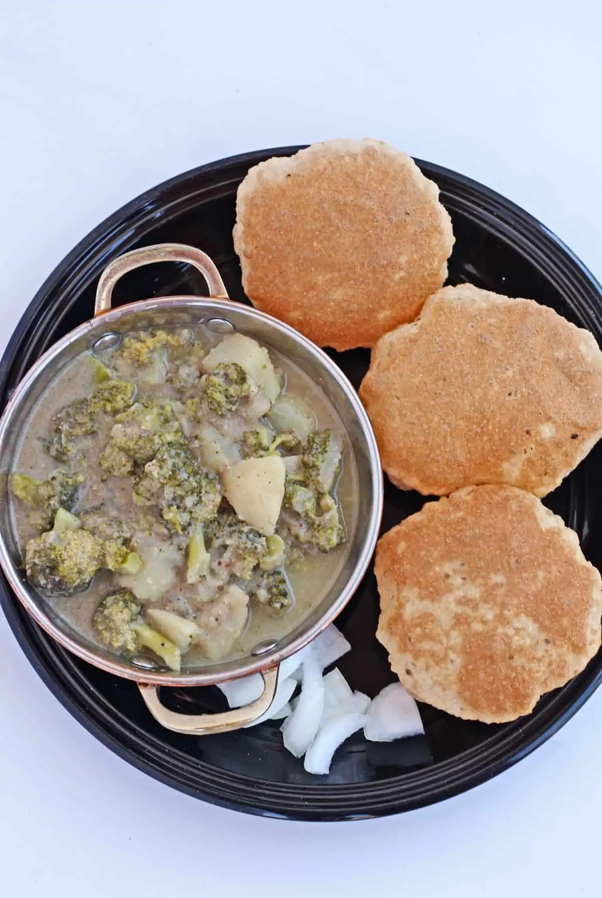 Broccoli and potato kurma with poori and onion pieces in a black playe