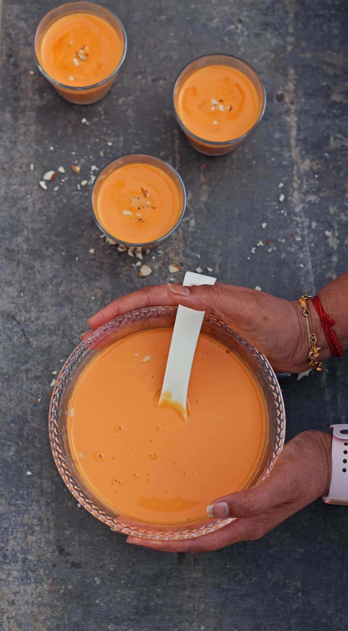 hands holding a bowl of carrot payasam and 3 glasses of kheer in the background.