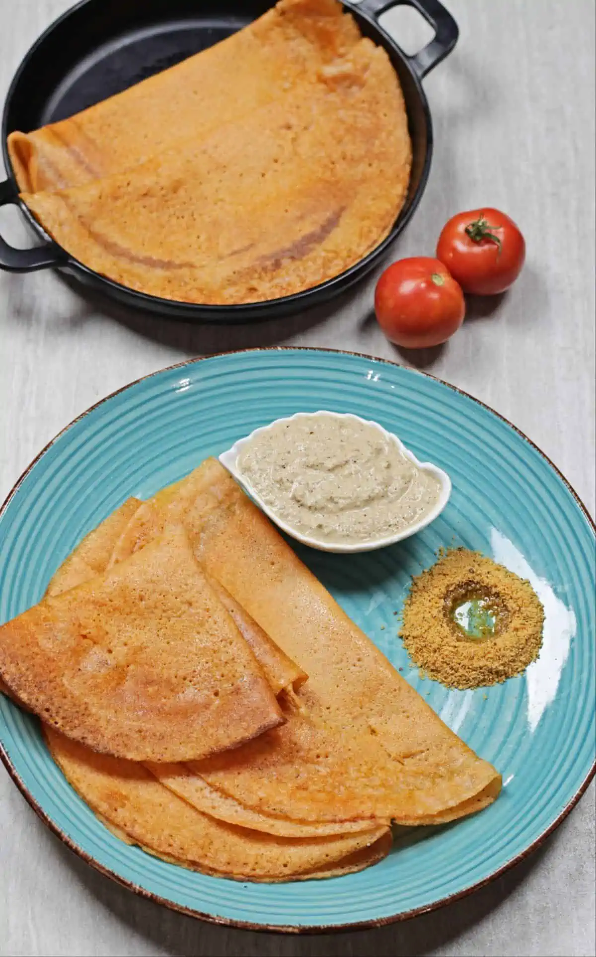 tomato dosa with chutney on a blue plate and black plate with tomatoes on side
