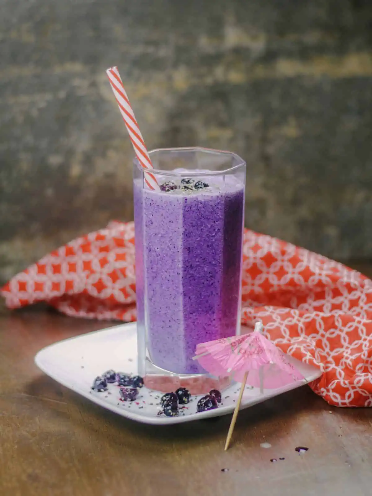Banana blueberry smoothie in a tall glass with straw and a paper umbrella on the side.